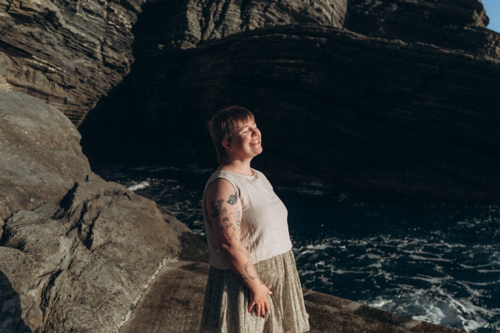 Katherine DeCoste is standing in front of an ocean cave and smiling into the sun.
